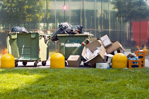 A row of three large green waste bins positioned on a paved surface in front of a glass building reflecting trees and a red object in the background. The left bin has a black bag draped over its lid, while the middle bin has a dark blue cover and appears to contain additional waste. To the right, there are several open cardboard boxes filled with miscellaneous rubbish, including paper, plastic, and packaging materials, some of which are leaning against the bins. Bright yellow plastic bollards are situated in front of the waste area, separating it from a well-maintained grassy verge. The scene suggests a typical waste disposal setup outside a commercial or residential building in Fulham, where Fulham House Clearance provides rubbish removal services, with the environment showing public space clearance activity under daylight conditions.