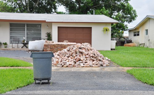 A pile of light pink and beige bricks is situated in the middle of a residential driveway in front of a single-story house with a beige exterior and a brown garage door. The bricks are stacked loosely, with some scattered around the base. To the left of the bricks, a large grey wheeled rubbish bin is positioned on a freshly paved black driveway. Behind the pile, the house features large windows with white frames and a small window box with greenery near the front door, visible on the left. To the right, part of a neighboring single-story house with a white exterior and a small exterior step can be seen. The scene is set in a suburban neighbourhood with well-maintained green lawns on either side of the driveway, and trees and foliage are visible in the background, indicating a bright, clear day. This image illustrates typical rubbish removal activity, possibly by Fulham House Clearance, in a residential area within the London borough of Fulham, relating to general rubbish disposal services.