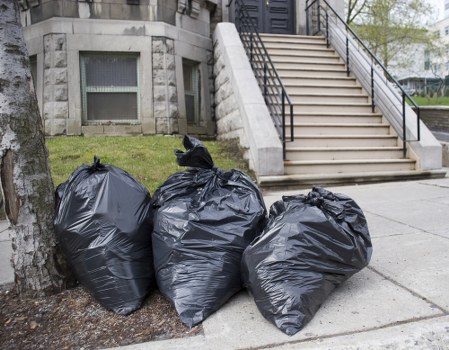 Three large black plastic rubbish bags filled with waste are placed on the pavement just beside a tree, in front of a stone building with a window. The bags are tightly tied at the top and appear to contain household rubbish or debris, ready for collection. Behind the bags, a staircase with metal handrails leads up to an entrance, and a neatly maintained grassy area surrounds the building. The pavement is concrete, and natural daylight illuminates the scene, suggesting an outdoor setting on a residential street possibly within Fulham, London, where Fulham House Clearance offers rubbish removal services. The scene is typical of a domestic rubbish clearance involving the disposal of accumulated waste, with no other objects or vehicles visible. This image accurately depicts a straightforward rubbish disposal situation in an urban environment, emphasizing the need for reliable waste collection services such as those provided by Fulham House Clearance.