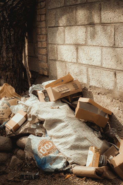 A narrow urban alleyway cluttered with a large, wheeled waste container in the foreground, which appears to be made of durable grey fabric or tarp material, with visible creases and signs of use. Behi
